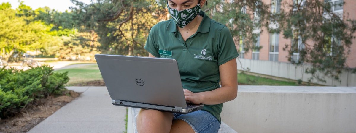 Student sitting outside with a laptop wearing a mask